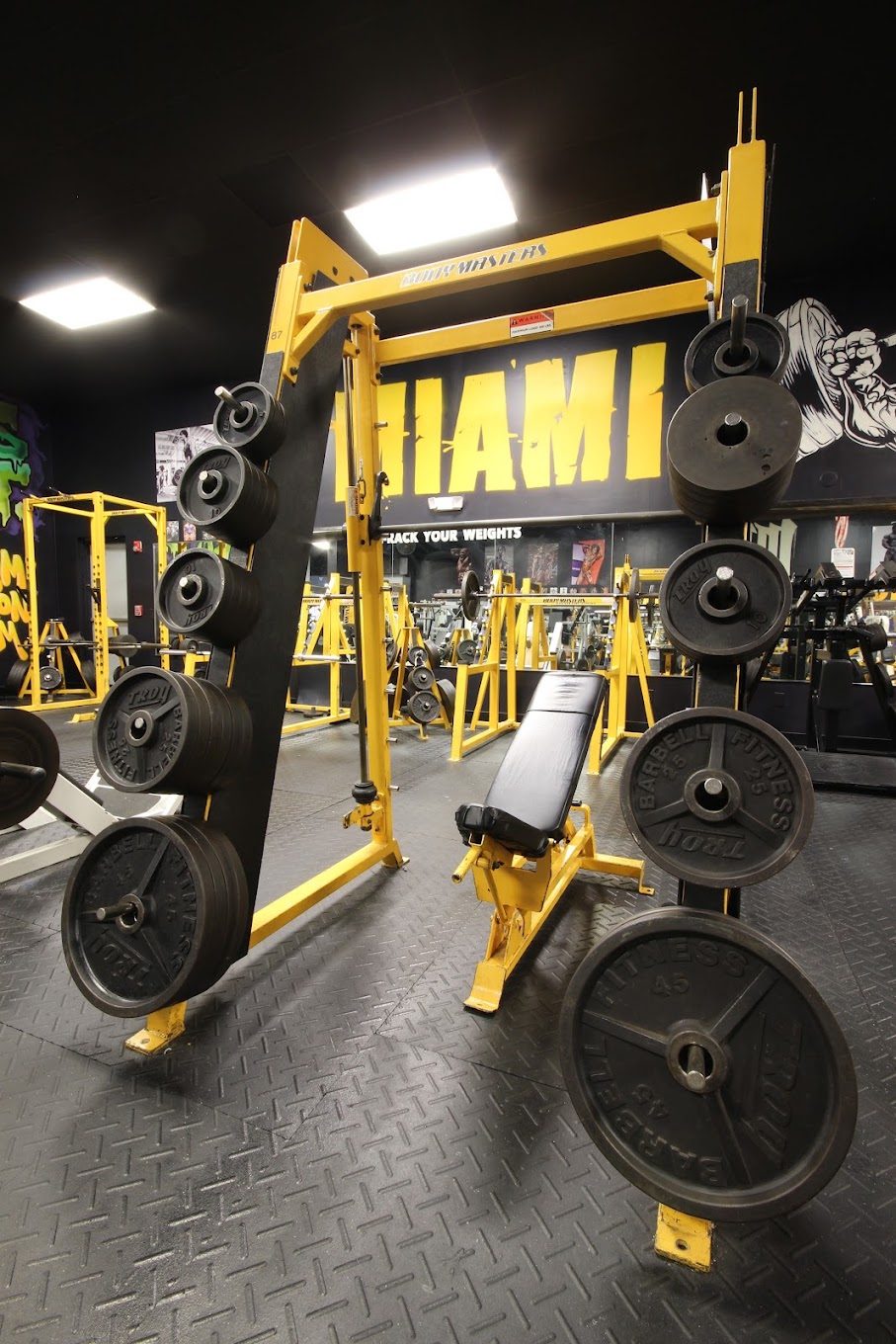 Miami Iron Gym equipment floor — yellow squat rack with weights and flat bench
