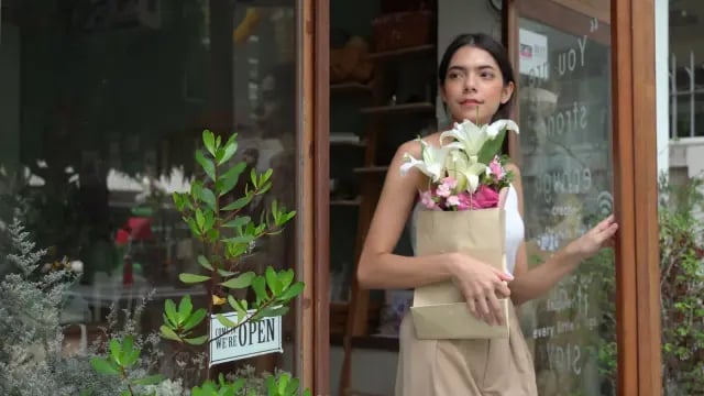 A shop owner at the door of her flower shop, holding a bouquet, with the open sign in the window